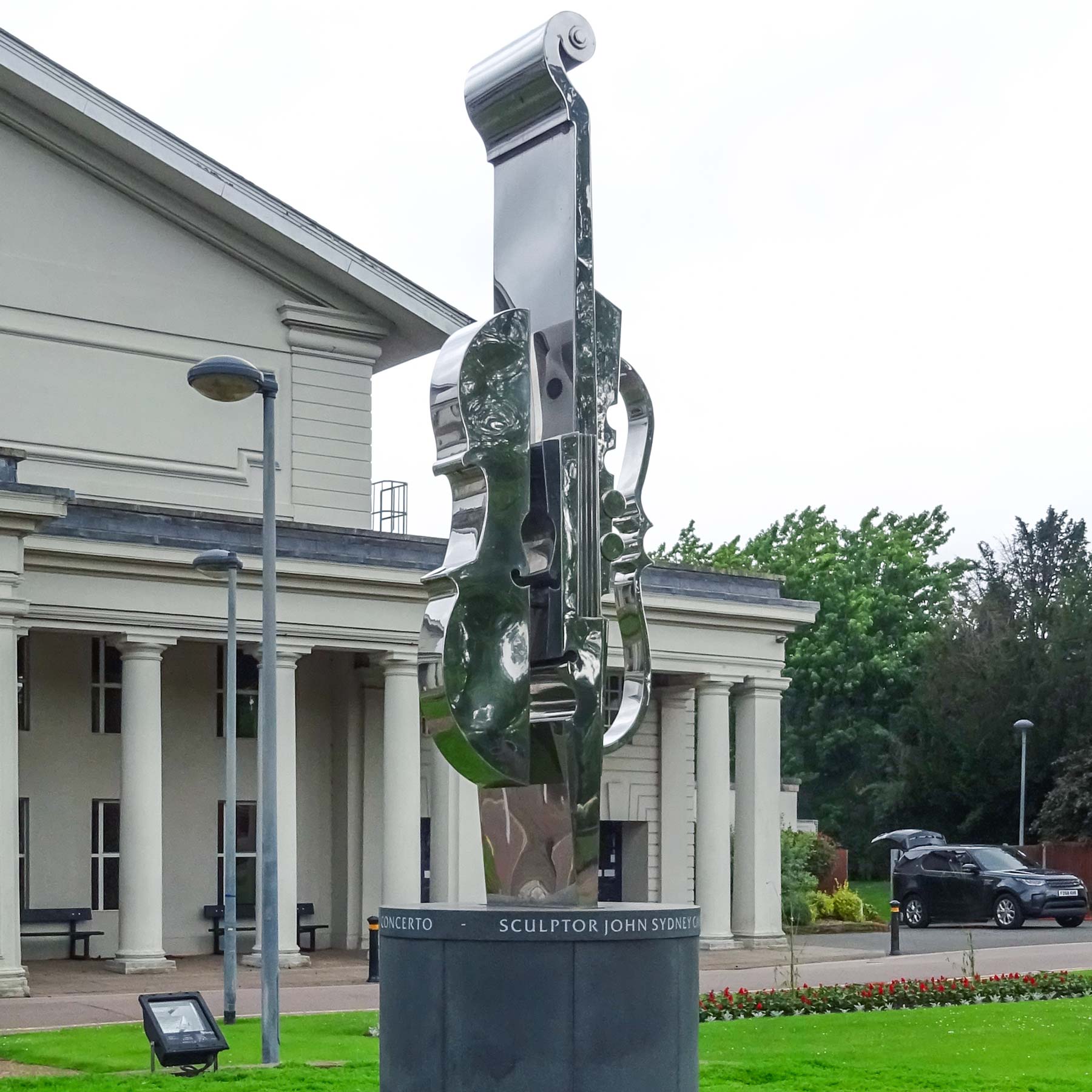 Photograph of the Concerto sculpture (resembles a violin form) in stainless steel, behind the sculpture is the triangle De Montfort Hall entrance and building