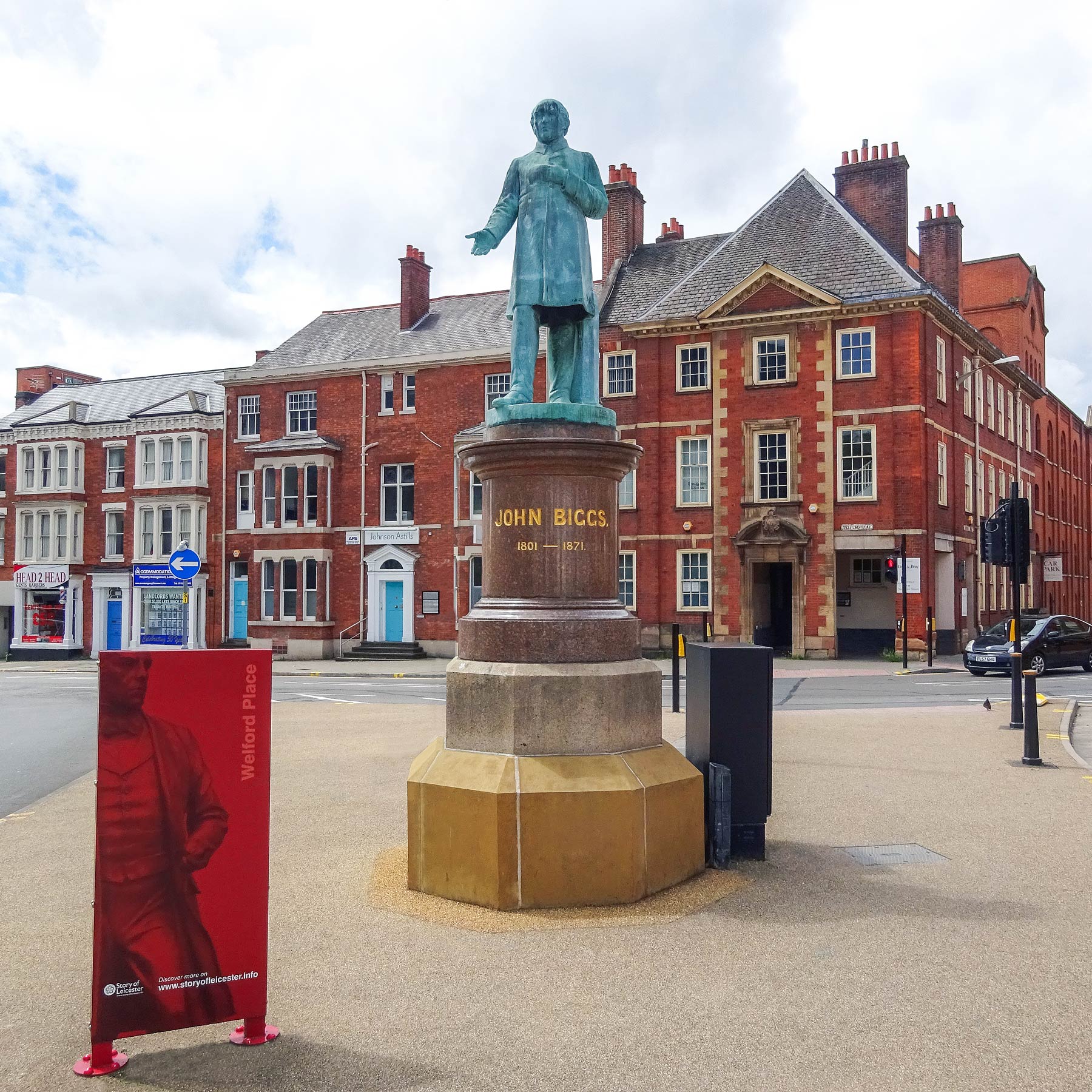 Photograph of the John Biggs statue green tinged, in the middle with one arm slighty out and hand open, on Welford Place a few metres past the end of Lower New Walk